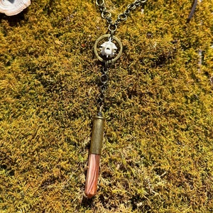 May include: A unique necklace featuring a pendant with a white bone-like element within a gold-colored ring, suspended from a dark chain and suede cord. The pendant is paired with a striped, reddish-brown crystal. The necklace rests on a bed of green moss.