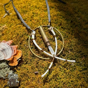 May include: A handcrafted necklace featuring a brass bullet casing pendant, accented with white bone-like elements and wrapped with copper wire. The necklace hangs on a dark gray leather cord, set against a backdrop of vibrant green moss.