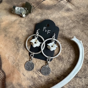 May include: Silver hoop earrings with a white bone charm and a silver coin pendant. The earrings are on a brown textured surface. The text "F+F" is visible on a black tag.