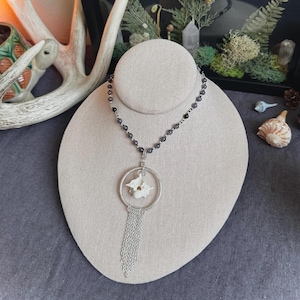 May include: A silver necklace with a black beaded chain and a pendant featuring a white flower design inside a circle with a silver tassel.