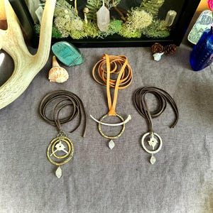 May include: Three leather cord necklaces with unique pendant designs. Each necklace features a different pendant with a bone and metal design. The pendants are all different shapes and sizes. The necklaces are all brown and are laid out on a brown surface.