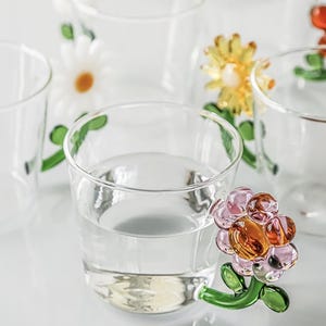 May include: Clear glass drinking glasses with colorful, decorative flower handles. The flowers are pink, orange, and yellow with green stems and leaves. The glasses are filled with a clear liquid.