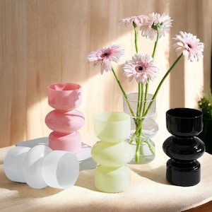 May include: Four glass vases in different colors, pink, white, light green, and black. Each vase has a unique, rounded, three-tiered design. The vases are arranged on a white surface with a book in the background. A bouquet of pink daisies is in the clear glass vase.
