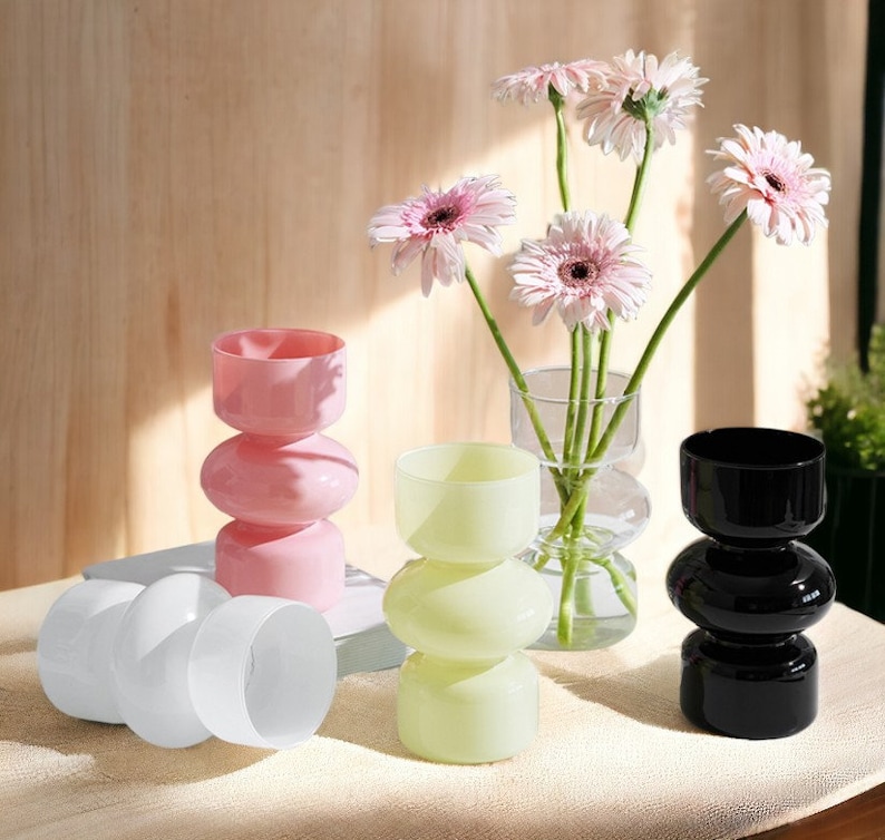 Peut inclure: Quatre vases en verre de diff&eacute;rentes couleurs&nbsp;: rose, blanc, vert clair et noir. Chaque vase a un design unique &agrave; trois niveaux arrondis. Les vases sont dispos&eacute;s sur une surface blanche avec un livre en arri&egrave;re-plan. Un bouquet de marguerites roses est dans le vase en verre transparent.
