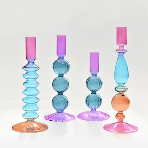 May include: Four colorful glass candle holders in shades of blue, pink, purple, and orange. The candle holders have unique designs, including stacked orbs, fluted bases, and cylindrical tops. The varying heights and shapes create a decorative display.