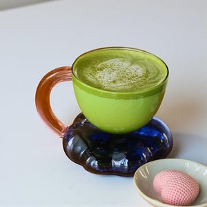 Könnte beinhalten: Eine Glastasse gefüllt mit grünem Matcha Latte, auf einer blau-violetten Glasuntertasse. Die Tasse hat einen transparenten Glaskörper mit grünem Rand und einem pfirsichfarbenen Henkel. Eine kleine weiße Schale mit einem rosa Mochi befindet sich in der Nähe.