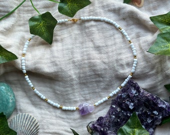 Könnte beinhalten: Eine weiße Perlenkette mit einem goldenen Verschluss und einem Anhänger aus violettem Amethystkristall.