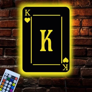 Puede incluir: Una luz LED negra con forma de carta de juego, el rey de corazones, con un borde amarillo y una «K» amarilla en el centro. La carta está montada en una pared de ladrillos. Un mando a distancia está en la esquina inferior izquierda.