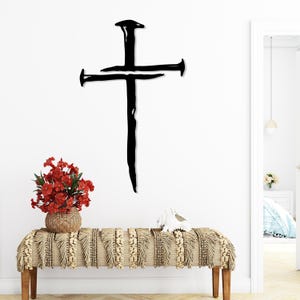 Puede incluir: Una gran cruz de metal negro hecha de clavos está montada en una pared blanca. Debajo, un banco con una cubierta tejida y con flecos sostiene un jarrón con flores rojas y un objeto decorativo blanco. La escena está bien iluminada.
