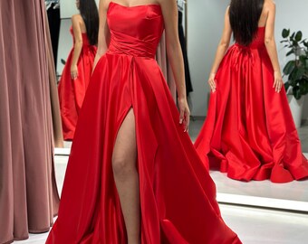 May include: A red strapless satin gown with a thigh-high slit and a fitted bodice. The gown has a flowing skirt and a simple design.