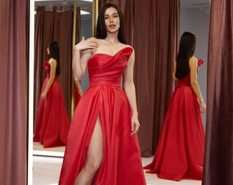 May include: A woman wearing a red strapless gown with a thigh-high slit. The gown has a fitted bodice and a flowing skirt. The woman is standing in a dressing room with a mirror behind her.