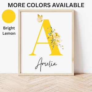 May include: Framed art print featuring a large yellow letter "A" with floral and butterfly accents. The name "Amelia" is written in cursive below the letter. The words "MORE COLORS AVAILABLE" and "Bright Lemon" are also visible.