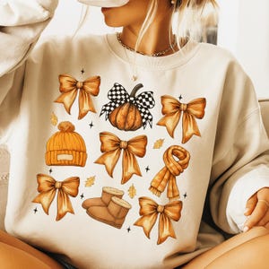 May include: Cream-colored sweatshirt featuring a fall-themed graphic. The design includes orange bows, a pumpkin with a checkered bow, a beanie, a scarf, and boots. The graphic is accented with small stars and leaves.