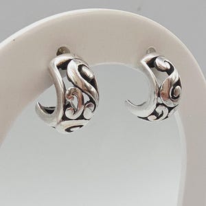 May include: A pair of silver-toned hoop earrings. Each earring has a crescent shape with an open filigree design featuring swirling patterns. The earrings are displayed on a white surface.