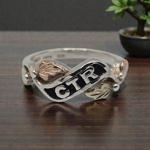 Puede incluir: Un anillo de plata con una banda central que presenta las letras CTR en negrita y relleno negro. El anillo está decorado con dos pequeños diseños de hojas texturizadas en color oro rosa a ambos lados sobre un acabado de plata pulida.
