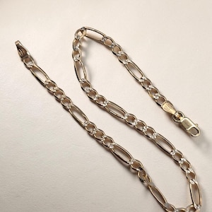 Könnte beinhalten: Ein goldfarbenes Figaro-Kettenarmband. Das Armband weist ein Muster aus länglichen ovalen Gliedern auf, die sich mit kleineren, kreisförmigen Gliedern abwechseln. Der Verschluss ist am Ende sichtbar. Das Armband wird vor einem neutralen Hintergrund präsentiert.