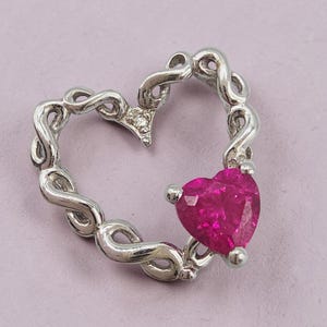 May include: A silver-toned pendant shaped like an open heart with a twisted metal frame. A single, faceted, vibrant pink heart-shaped gemstone is set into the lower right side of the frame, creating a contrast with the polished metallic surface.