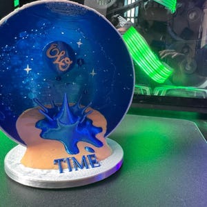May include: A 3D-printed sculpture featuring a blue and gold design with the text "TIME". The sculpture has a celestial theme with stars and a splash-like element. The letters "ELO" are also visible on the sculpture.
