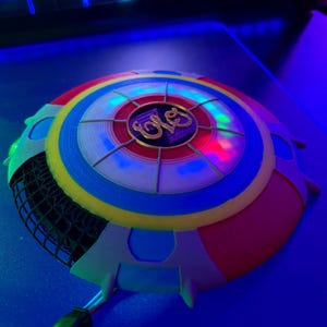 Puede incluir: Objeto circular multicolor con un logotipo central y luces. El objeto presenta secciones rojas, amarillas, azules y blancas, con una zona de malla negra. El logotipo central es dorado. Las luces son de varios colores.