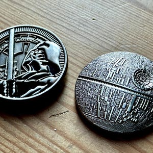 May include: Two silver metal coins. One coin features Darth Vader with a lightsaber and the text "HUGO VADER.COM". The other coin depicts the Death Star. Both coins are round and have detailed designs.