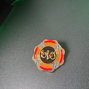 Könnte beinhalten: Goldfarbene Anstecknadel mit den Buchstaben "ELO" in einem stilisierten Schriftzug, vor schwarzem Hintergrund in einem kreisförmigen Design. Die Anstecknadel hat einen dekorativen, mehrschichtigen Rand mit roten, orangefarbenen und goldenen Akzenten. Wahrscheinlich ein Sammlerstück.