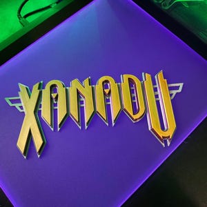 May include: A purple surface featuring the gold word "XANADU" in a stylized font. The letters have a metallic, three-dimensional effect. The background is a darker shade of purple, with green and black accents.