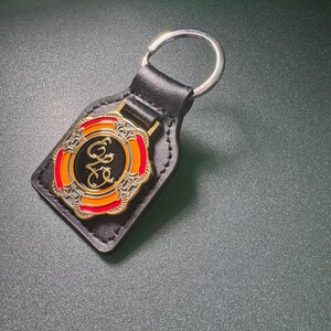 May include: Black leather keychain with a silver key ring. The keychain features a decorative emblem with orange, red, and gold accents. The emblem has the letters "E" and "G" in a stylized font.