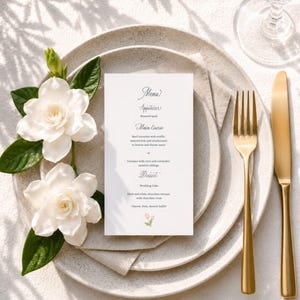 Puede incluir: Una tarjeta de menú rectangular blanca con texto negro que enumera platos como codorniz asada, solomillo de ternera y pastel de bodas, colocada sobre un plato de cerámica moteada con dos flores de gardenia blancas, un tenedor y un cuchillo dorados.