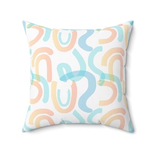 Puede incluir: Una almohada blanca con un patrón abstracto colorido de arcos azul pastel, melocotón y amarillo.