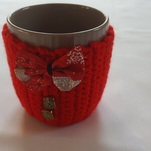 Peut inclure: Un manchon de tasse en crochet rouge avec un nœud en tissu et deux boutons dorés. Le manchon est sur une tasse en céramique brune.
