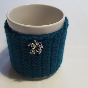 Peut inclure: Un manchon de tasse en tricot bleu sarcelle avec un charme en feuille d'argent. Le manchon est fait d'un fil doux et texturé et est conçu pour s'adapter à une tasse de taille standard.
