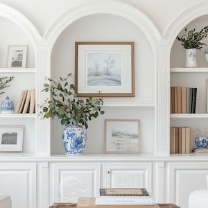Puede incluir: Estanterías empotradas blancas con arcos superiores, que exhiben libros, arte enmarcados y jarrones decorativos azules y blancos. Los estantes están llenos de libros de tonos neutros, vegetación y arte paisajístico enmarcado. Dos sofás blancos con cojines azules estampados están en primer plano.