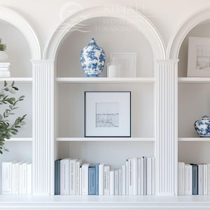 May include: A built-in white bookshelf with arched openings, displaying decorative items. The shelves hold stacks of books, framed artwork, and blue and white porcelain vases. A vase with greenery adds a natural element to the display.