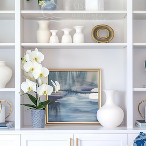 May include: White built-in shelving unit with various decorative items. Includes white vases, a framed landscape painting, gold-toned sculptures, potted plants, and a potted orchid with white flowers. The shelves are filled with home decor.