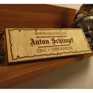 May include: A rectangular wooden nameplate with the name "Anton Schimpf" and the title "CNC - OPERATOR" engraved in a decorative font. The website address "WWW.GRAINLY.DE" is also visible. The nameplate is light brown and has a decorative border.