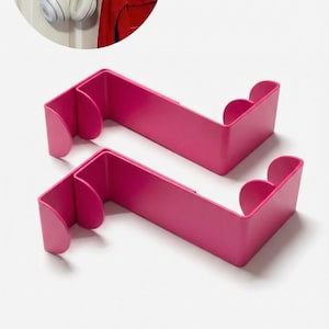May include: Two bright pink over-the-door hooks. The hooks are designed to hang over a door and have a curved shape. The image also shows a pair of white headphones and a red backpack hanging on a door using similar hooks.