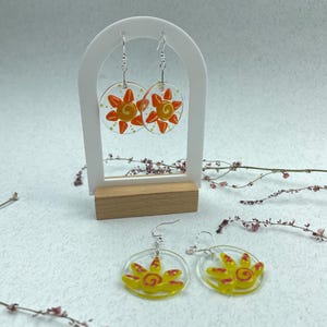 Puede incluir: Pendientes de acrílico transparente con diseño de sol. Los pendientes tienen forma circular con un diseño de sol amarillo y detalles en rojo. Los pendientes se exhiben en un soporte blanco con base de madera.