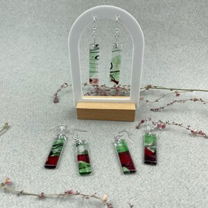 Puede incluir: Pendientes colgantes rectangulares con un diseño abstracto rojo, verde y blanco. Los pendientes se exhiben en un soporte blanco con una base de madera, y también se colocan sobre una superficie gris. Los pendientes tienen ganchos plateados.