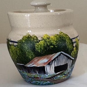 May include: A beige ceramic jar with a lid, hand-painted with a rural landscape scene. The artwork depicts a white barn with a rusty roof, green trees, and blue and white flowers. The jar has a rounded shape.