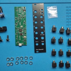 May include: An assortment of electronic components, including a green circuit board, black panel with circular holes, and various switches and connectors. The panel has the text "Thresh", "CV scale", "CV in", "Clock", and "Timo Rozendal".