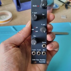 Puede incluir: Un módulo electrónico negro con perillas y conectores, etiquetado como "OAM Time Machine Expander". Cuenta con controles para "Mode", "F start", "Set", "F end" y "Res". El módulo también tiene indicadores "LPF", "BPF" y "HPF", junto con conectores "Stereo".