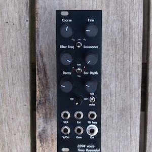 May include: Black modular synthesizer module with knobs and input/output jacks. The module has text labels including "Coarse," "Fine," "Filter Freq," and "Resonance." The text "3394 voice Timo Rozendal" is printed at the bottom. The module is on a wooden surface.