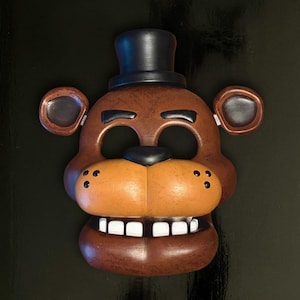 Puede incluir: Máscara marrón de Freddy Fazbear con sombrero de copa negro, nariz negra y dientes blancos. La máscara tiene aberturas para los ojos y la boca, y pequeños puntos negros en las mejillas. Ideal para cosplay o fiestas de disfraces.