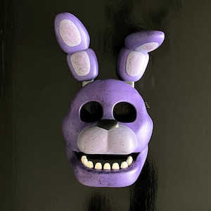 May include: Purple and white plastic mask of a cartoon bunny with black eyes and a toothy grin. The mask has a textured surface and is designed to look like a character from a video game.