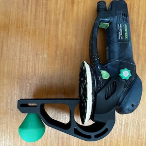 Op de afbeelding: Een zwarte en groene elektrische schuurmachine met een groene schuurpad en een zwarte standaard met een groene bal. De schuurmachine heeft de tekst "FESTOOL" en "RO 150 FEQ" erop.