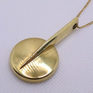 Op de afbeelding: Een goudkleurige metalen hanger aan een ketting met een cirkelvormige schijfbasis en een taps toelopende, langwerpige driehoekige arm die vanuit het midden uitsteekt. De hanger hangt aan een fijne goudkleurige ketting met een gladde, gepolijste afwerking.