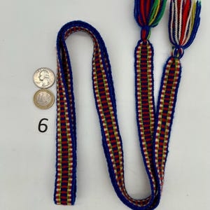 Pu&ograve; includere: Un cinturino blu intrecciato con strisce rosse, gialle e bianche e nappe alle estremit&agrave;. Le nappe sono fatte di fili colorati. Il numero 6 &egrave; stampato sulla superficie. Sono visibili anche due monete.