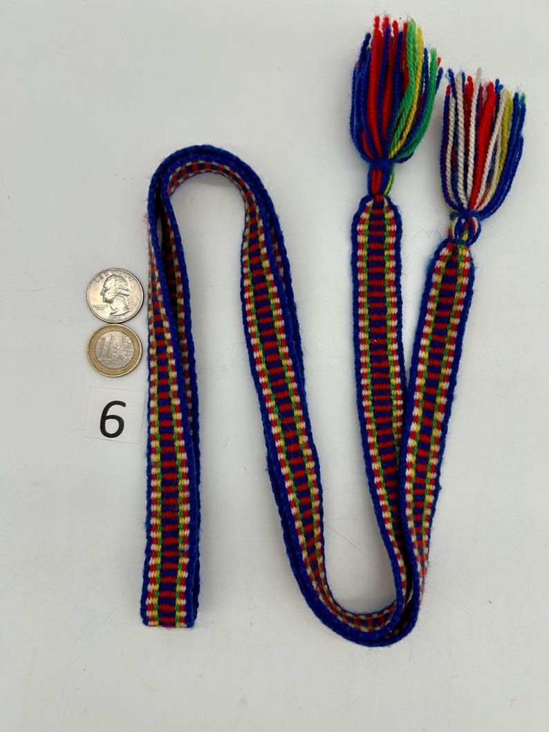 Pu&ograve; includere: Un cinturino blu intrecciato con strisce rosse, gialle e bianche e nappe alle estremit&agrave;. Le nappe sono fatte di fili colorati. Il numero 6 &egrave; stampato sulla superficie. Sono visibili anche due monete.