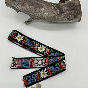 May include: Black ribbon with colourful floral embroidery featuring white, red, and blue flowers. The ribbon is displayed with a piece of driftwood and two coins. The ribbon's design is reminiscent of traditional folk art.