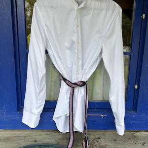May include: A crisp white long-sleeved button-down shirt featuring a classic collar and cuffs. A long, narrow woven belt in black, pink, and white with multi-colored tasseled ends is tied around the waist of the shirt, creating a gathered effect.